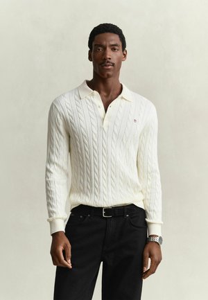 Homme portant un polo blanc en maille torsadée à manches longues, un pantalon noir et une montre-bracelet argentée, debout devant un fond uni.