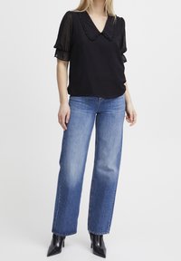 Sort bluse med gennemsigtige, flæsebesatte korte ærmer og V-hals; stylet med blå høj talje lige ben jeans og sorte spidse hæle.