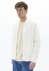 Chaqueta bomber blanca de tela suave, con cierre de cremallera y bolsillos laterales, llevada sobre una camisa beige clara, combinada con jeans de mezclilla azul.