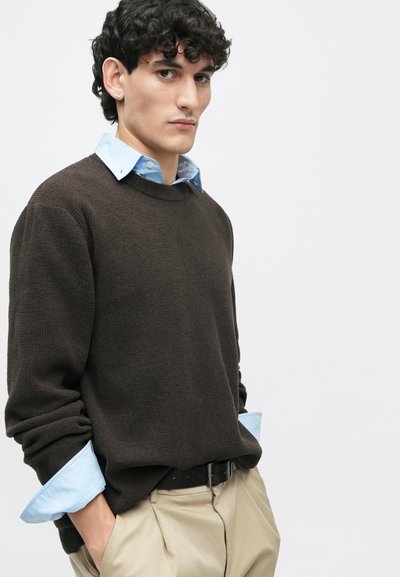Rudas tekstūruotas megztinis su apvalia iškirpte ir sulankstytais šviesiai mėlynais marškinių rankovėmis, derinamas su smėlio spalvos kelnėmis ir juodu diržu.