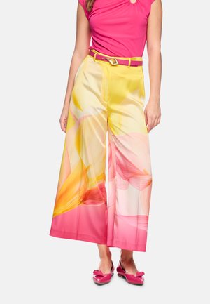 Frau trägt ein pinkes Top mit gelb-pinken, weit geschnittenen Blumenhosen, einen pinken Gürtel mit goldener Schnalle und pinke spitze Ballerinas mit Schleifen.