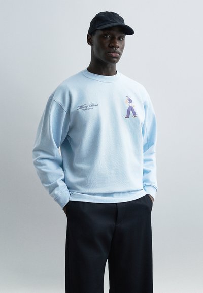 Jeune homme portant un sweat-shirt bleu clair avec une illustration d'un homme qui marche, un pantalon noir et une casquette foncée, debout les mains dans les poches.