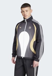 Melnas jakas ar baltu un zeltītu spirālveida dizainu, augstu apkakli, pilnu priekšējo rāvējslēdzēju un baltām Adidas svītrām uz piedurknēm; savienota ar melnām biksēm.