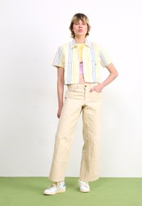 Beskaaret gul og hvid stribet skjorte, lyse lilla tanktop, beige bukser med vidde og hvide sneakers med blå detaljer, iført mod en hvid baggrund.