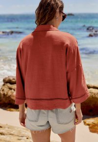 Veste en lin terracotta à coupe décontractée, manches trois-quarts, et couture décorative sur l'ourlet, associée à un short en denim léger.