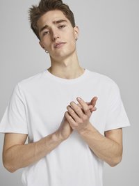 Camiseta blanca de cuello redondo hecha de algodón suave, con mangas cortas. El modelo muestra las manos juntas frente a un fondo gris neutro.