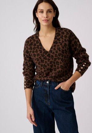 Vrouw draagt een bruine trui met luipaardprint en gouden knopen op de mouwen en hoge taille donkerblauwe spijkerbroek, hand in de zak, neutrale achtergrond.
