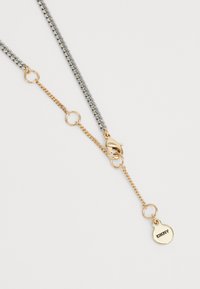 Collier à maillon bicolore avec fermoir en or en forme de homard et petite étiquette ronde en or gravée "DKNY" sur fond blanc.