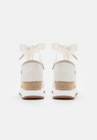 White shoes with a textured espadrille wedge heel, featuring an ankle tie with cream ribbon and metallic accents on the ties.
