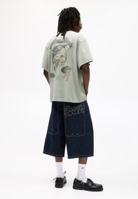 Tricou oversized de culoare verde deschis cu imprimeu grafic pe spate, asociat cu culottes din denim închis, ce prezintă cusături vizibile și branding. Pantofi negri.