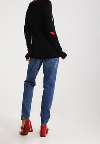 Czarny dziergany sweter z czerwonymi grafikami ust i oczu na rękawach, w połączeniu z niebieskimi jeansami o prostych nogawkach oraz czerwonymi butami na słupku.