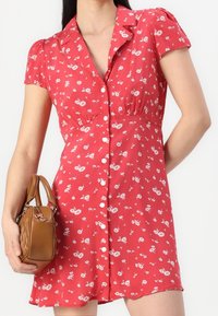 Rotes Blumenkleid mit kurzen Puffärmeln, knopflosem Vorderteil, tailliertem Schnitt und ausgestelltem Rock. Kombiniert mit einer kleinen braunen Handtasche.