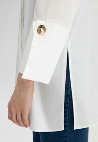 Witte shirt met een manchetknoop van rond goud, een zijsplit en contrasterende blauwe denim eronder. Glad textuur en op maat gemaakte ontwerp.