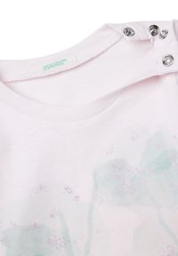 Zbliżenie na dekolt jasnoróżowej koszulki dla niemowląt z trzema metalowymi zatrzaskami, wyblakłym kwiatowym nadrukiem oraz metką United Colors of Benetton.