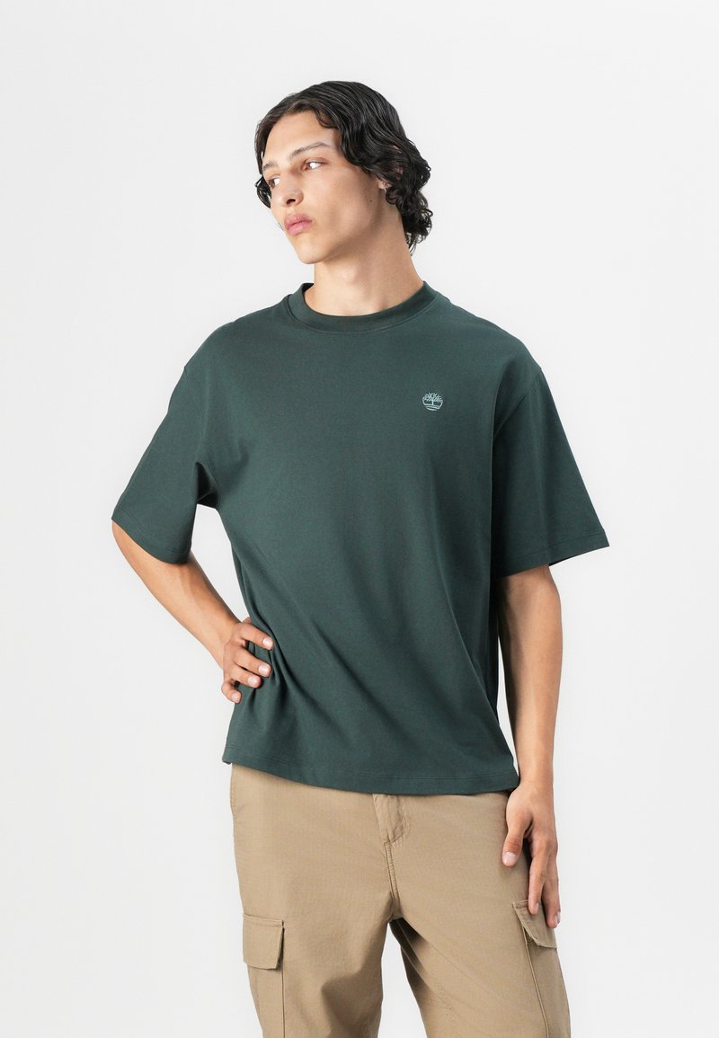 Camiseta de manga corta verde oscura de algodón, con cuello redondo y un pequeño logo en el pecho. Combinada con pantalones cargo beige.