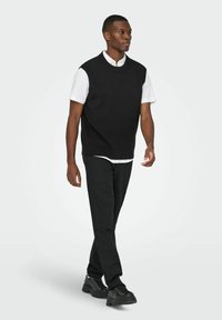 Schwarzer ärmelloser Pullover über einem weißen Kragenhemd, kombiniert mit schwarzen Hosen und klobigen schwarzen Schuhen, was einen geschichteten monochromen Look kreiert.