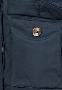 Sección de chaqueta de tela azul marino que cuenta con un parche de bolsillo prominente con un botón metálico y un pequeño detalle en forma de lazo sobre el bolsillo.