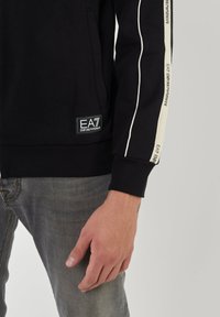 Felpa nera con strisce bianche sui lati, con un patch del logo all'orlo, realizzata in materiale morbido. Jeans grigi visibili nella parte inferiore.