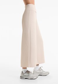 Beige wijde broek met een hoge tailleband, geribbelde textuur en een losse pasvorm, gecombineerd met lichtgrijze sneakers en witte enkelsokken.