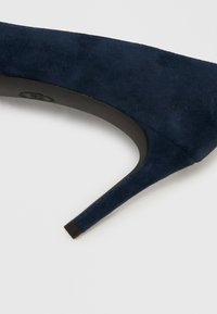 Temno modra semišasta salonarka z visokim petom, z zašiljenim prstom in elegantnim črnim podplatom, z teksturirano zgornjostjo in tankim dizajnom pete.