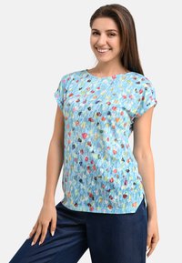 Blaues Kurzarm-T-Shirt mit einem bunten Punktmuster. Leichter Stoff, Rundhalsausschnitt, Seitenschlitze, kombiniert mit dunkelblauen Hosen.