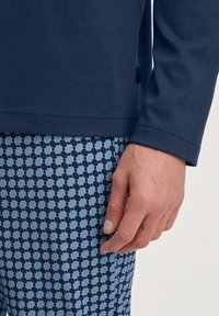Marineblauwe langemouwen shirt boven een patroon van blauwe en zwarte broek met hexagonale ontwerpen. Glad materiaal, close-up van arm en hand.