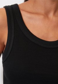 Detailní záběr na osobu v černém žebrovaném tílku, ukazující detaily výstřihu a ramen na světlé pleti.