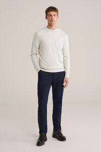 Ljusbeige tröja med rund halsringning, kombinerad med marinblå chinos. Modellen står i svarta skor och visar upp en avslappnad passform och enkel design.
