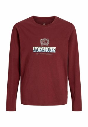 Chłopiec o kręconych włosach, ubrany w koszulę w kratę zapinaną na guziki, na czerwonym t-shircie z nadrukiem, czarne dżinsy i brązowe buty, stojący na jednolitym białym tle.