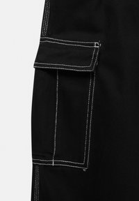 Detail černých cargo kalhot s nášivkou, bílé šití, které ukazuje pevnou tkaninu a strukturovaný design.