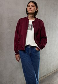 Chaqueta bomber burdeos con puños de rayas blancas, superpuesta a una camiseta blanca con un pequeño gráfico oscuro, combinada con jeans de pierna ancha azules.