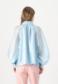 Blusa in satin azzurro chiaro con maniche a sbuffo e scollatura arricciata. Polsini con bottoni. Indossata con pantaloni rosa chiaro, che mostrano una texture liscia.