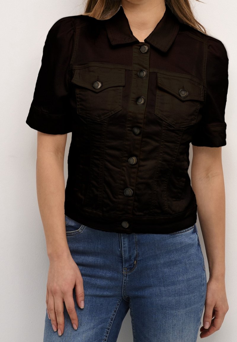 Zwarte korte mouw jacket met een knoopsluiting aan de voorkant, voorzien van twee borstzakken en een getailleerde pasvorm, in combinatie met lichtblauwe jeans.