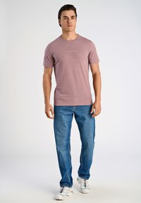 Kortärmad, lila t-shirt med rund halsringning och "LINDBERGH"-logotyp i ljus text, matchad med blå jeans och vita sportskor.