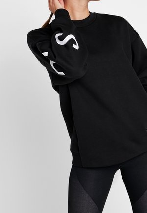 Kobieta ubrana w luźną czarną bluzę z białymi literami na rękawie i teksturowane czarne legginsy na jednolitym tle.