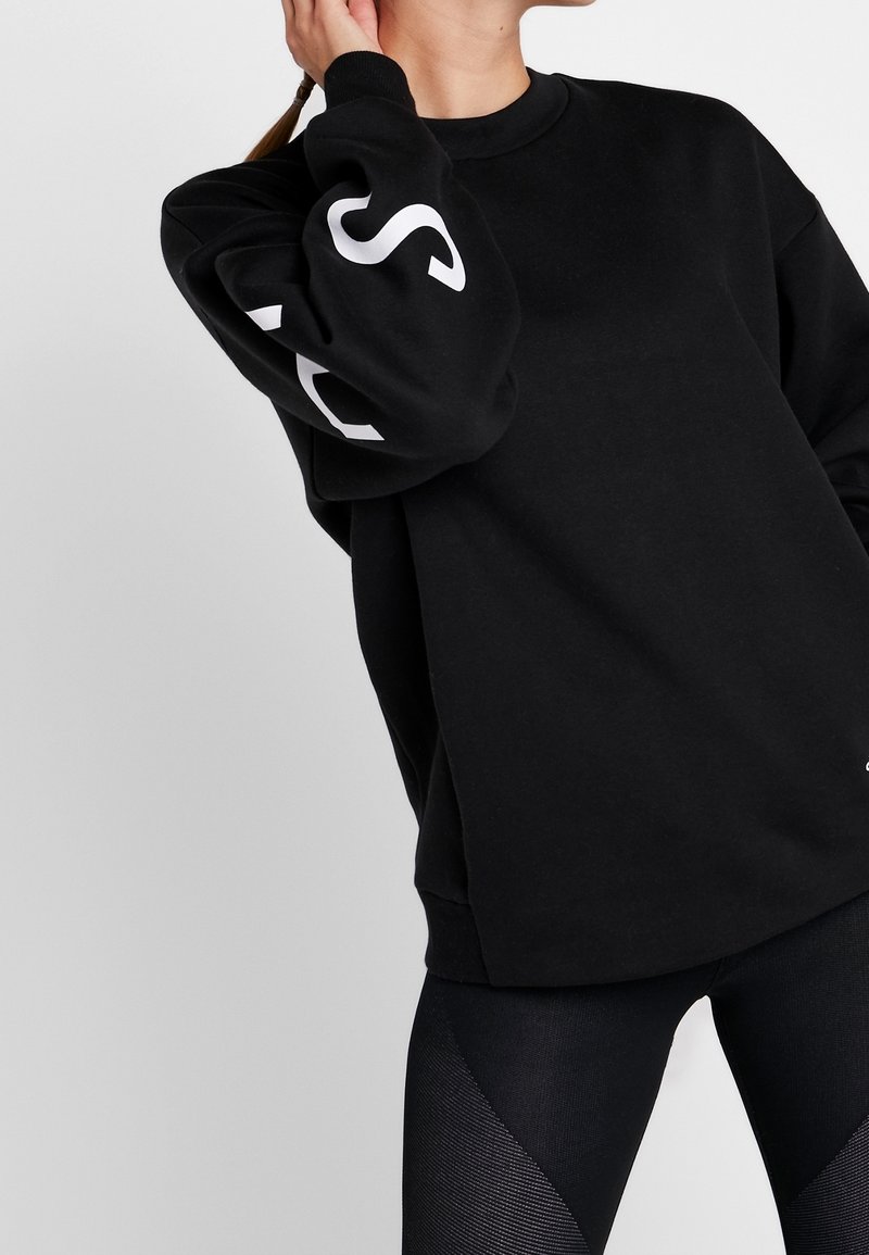 Femme portant un sweat noir ample avec des lettres blanches sur la manche et des leggings noirs texturés sur un fond uni.