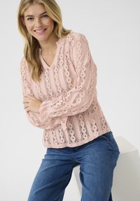 Roza čipkast bluzo z V-izrezom, dolgimi rokavi z elastičnimi manšetami in vzorci s perforacijami, kombinirana z modrimi kavbojkami. Gladka tekstura.