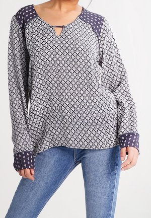 Bluse mit langen Ärmeln, die ein marineblau-weißes geometrisches Muster aufweist, einen V-Ausschnitt mit einem Schlüsselloch-Ausschnitt und kontrastierenden gemusterten Manschetten hat.