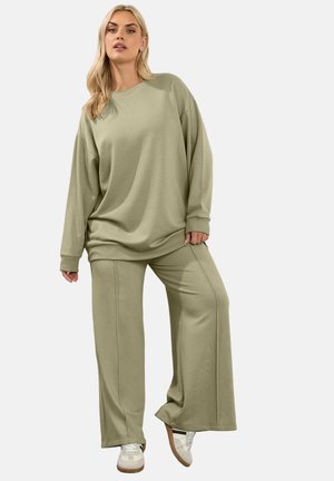 Femme blonde portant un sweat-shirt ample vert olive et un pantalon à jambes larges avec des baskets blanches, debout avec une jambe légèrement levée.