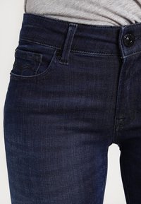 Jeans de mezclilla azul oscuro con una textura suave, diseño clásico de cinco bolsillos y cierre de botón en la parte frontal. Presenta detalles de costura sutiles.
