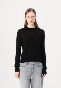 Svart ribbet strikkegenser med lange ermer, rund hals og en tettsittende silhuett, kombinert med lysegrå jeans.