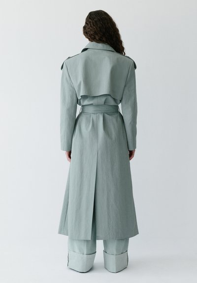 Kvinde med krøllet hår står med ryggen til iført en lys grå trenchcoat med bælte og bukser med vide ben mod en ensfarvet baggrund.