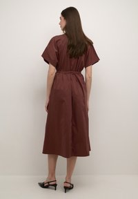 Brown, knee-length dress with short sleeves, cinched waist, and smooth texture. Paired with black slingback heels.