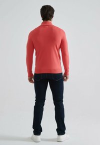 Pull-over en corail, texture côtelée, coupe slim, porté avec un jean bleu marine et des baskets blanches, présenté de dos sur un fond uni.