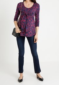 Laivastonsininen ja viininpunainen kukallinen äitiysfarkkupaita, joissa on 3/4-hihat, solmittu etuosan yksityiskohta, yhdistetty tummiin farkkuihin ja mustiin ballerinoihin, pitäen mustaa laukkua.