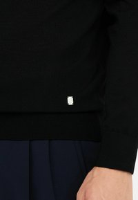 Maglione nero a maglia con orlo a coste; presenta una piccola etichetta logo argentata sul lato inferiore, abbinato a pantaloni plissettati blu navy.