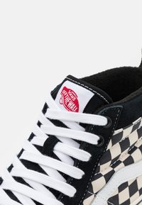 Mustra mustreeritud must ja kreemikollane kanvas toss valgete paeltega, pehme kraega ja mustade nubukaktsentidega. Omab punast Vans logo silti.
