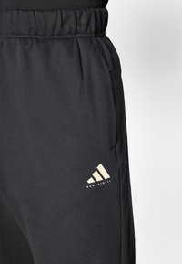 Schwarze Jogginghosen mit einem elastischen Bund, Seitentaschen und einem beigen Adidas-Logo mit einem Dreistreifen-Design an der unteren Beinseite.