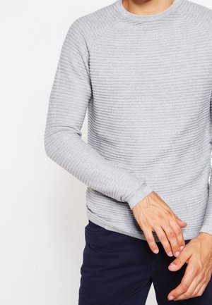 Mężczyzna w jasnoszarym prążkowanym swetrze z długim rękawem i ciemnoniebieskich spodniach, poprawiający rękaw rękami na tle białego tła.