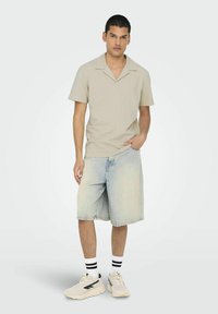 Polo beige a maniche corte con un motivo testurizzato, abbinato a pantaloni corti di jeans azzurri. Il modello indossa sneakers bianche con dettagli neri.